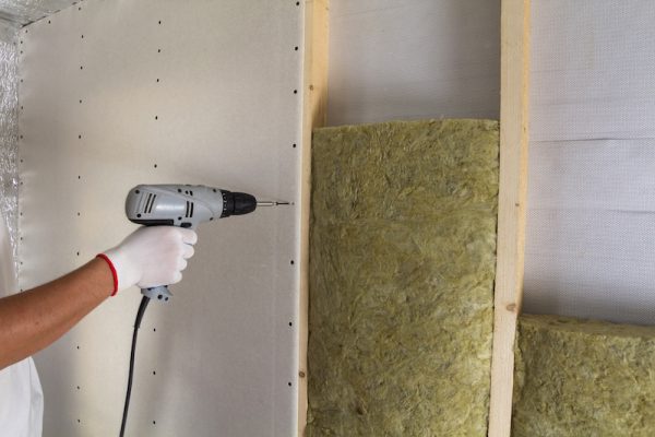 Close-up of worker hand with electric screwdriver fastening drywall to wooden timber frame with insulation mineral rock wool staff. Warm home, low energy bills, construction and renovation concept. Close-up of worker hand with electric screwdriver fastening drywall to wooden timber frame with insulation mineral rock wool staff. Warm home, low energy bills, construction and renovation concept.